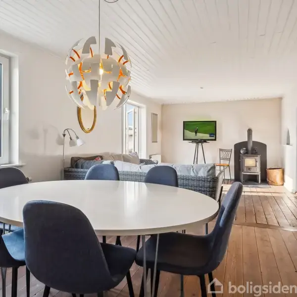 Rundt spisebord med seks blå stole står i lyst rum. En unik lysekrone hænger over bordet. Sofaen er placeret ved vinduet, med en brændeovn og fjernsyn ved siden af.