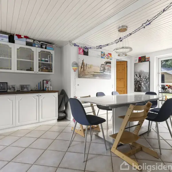 Et spisebord med stole omgivet af et lyst rum. Skabe står langs væggen med dekorationer ovenpå. En dør og glasdøre fører til en udendørs visning. Ingen tekst fundet.