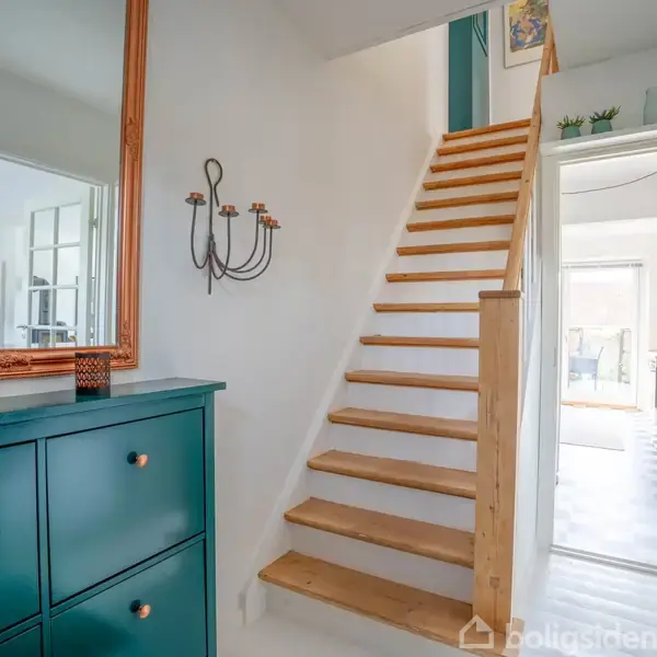 Trætrappe stiger op i en lyspåvirket gang, flankeret af en grøn kommode med spejl og væghængt lysestage til venstre, der leder til et lyst køkkenområde i baggrunden.
