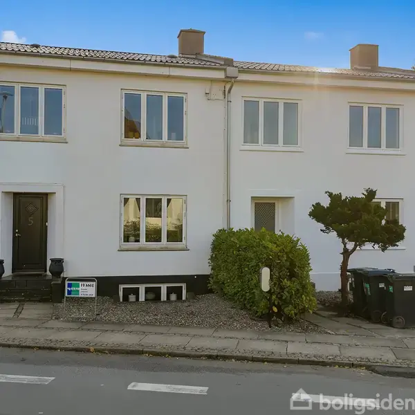 Et hvidt rækkehus med tre vinduer og en sort dør står stille ved en gade. Foran er en grøn busk og affaldscontainere. Skiltet viser "Til salg DK Boligsyd / Nybolig."