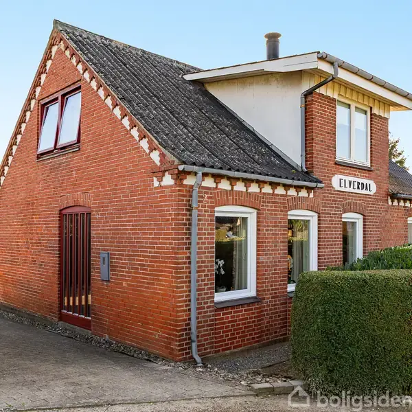 Rødt murstenshus står stille omgivet af hække og en indkørsel. "ELVEDAL" er skrevet over dørene; et træskur kan ses i baggrunden under en blå himmel.