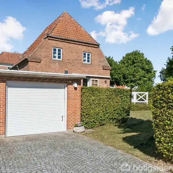Et rødt murstenshus med gavltag står ved siden af en garage med hvid port. Hæk omkranser græsplæne, og brosten dækker indkørsel. Skyfri blå himmel og grønne træer i baggrunden.