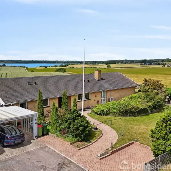 En bungalow med sort tag og rund indkørsel, omgivet af grønne buske. To biler holder i carporten. Landskabet er en åben mark med udsigt til en fjord i baggrunden.