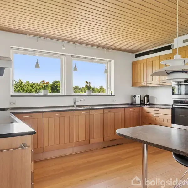 Køkken med træskabe og sorte bordplader, hvor en stor vask er installeret under et stort vindue. Hængelamper hænger over køkkenøen, og der er moderne hvidevarer mod højre.