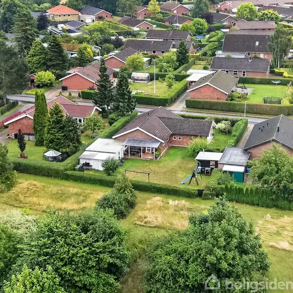 En villa med rød mursten og mørkt tag ligger i et grønt boligområde med flere lignende huse. Haven indeholder et legehus og gyngestativ, omgivet af træer og buske.
