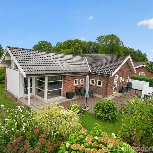 Et rødstenshus med gråt tag står midt i en frodig have med blomster og træer. Der er en terrasse foran huset, og flere omgivelser med grønne buske og hegn.