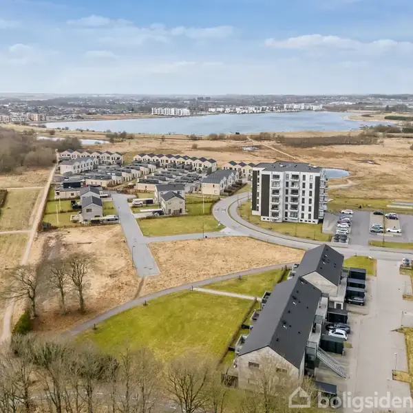 Moderne lejlighedskompleks står stille, omgivet af græsarealer og veje, med en sø i baggrunden under en blå himmel.