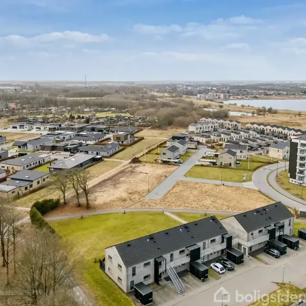 Boligblokke ligger stille i et nybyggerkvarter omgivet af græsarealer og træer. Flere rækker af moderne huse strækker sig ud mod en fjern sø, under en let overskyet himmel.
