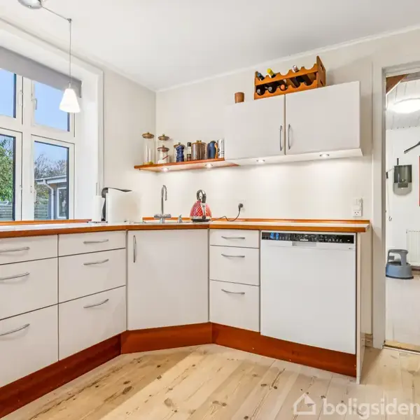 Køkken med hvide skabe og træbordplader, opvaskemaskine, og vask. To hængende lamper over vindue, udsigt til have. Åben dør fører til tilstødende rum med trægulv.