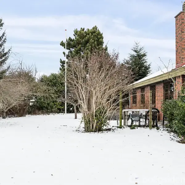 Et rødt murstenshus står ved siden af et snedækket område med et bord og stole. Træer og buske omkranser området under en klar himmel.