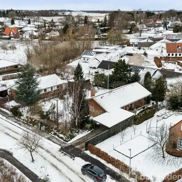 Snedækket boligområde med flere huse, træer og veje. Et hus og en bil på en vej er i fokus. Omgivelserne er dækket af sne, mens andre huse ses i baggrunden.
