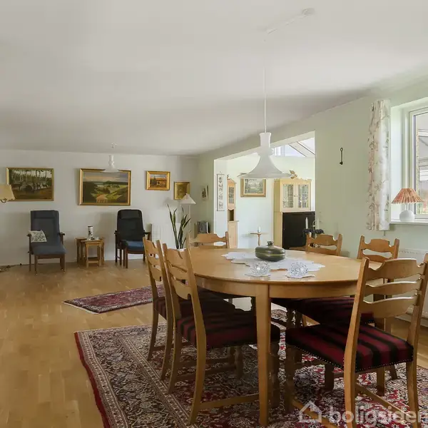 Et rundt spisebord med træstole er centralt placeret i en stue. Rummet har hvidmalede vægge, billeder og to lænestole bagerst, samt et vindue med gardiner til højre.