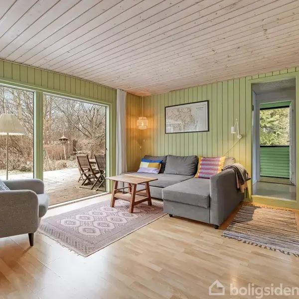 Stue med grå sofa ved siden af en lænestol, foran store glasdøre til en terrasse. Rummets vægge er lysegrønne, og der er et skrivebord i højre hjørne.