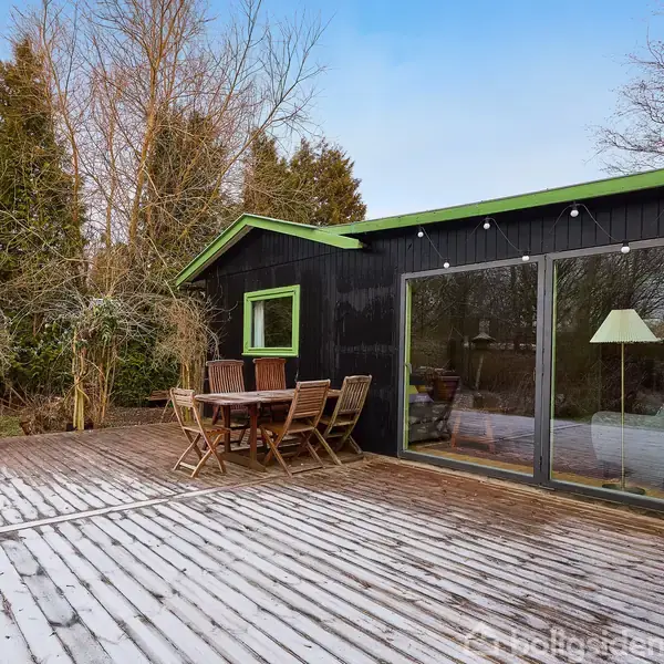 Et sort træhus står på en terrasse med bord og stole. Store glasvinduer viser en lampe indendørs. Omgivet af træer i en naturlig have.