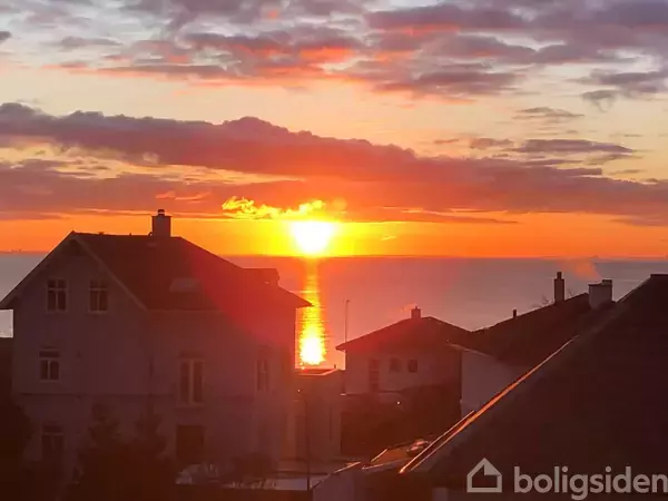Solnedgang skinner over havet bag hustage, hvor solens lys reflekteres i vandet. Skyerne er farvet orange og lilla, hvilket fremhæver den rolige aftenstemning.