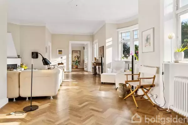 Stue med hvid sofa ved pejs, gulvlampen tændt, i lyst rum med parketgulv og store vinduer. Der er også en hvid stol og en trægulvstol.