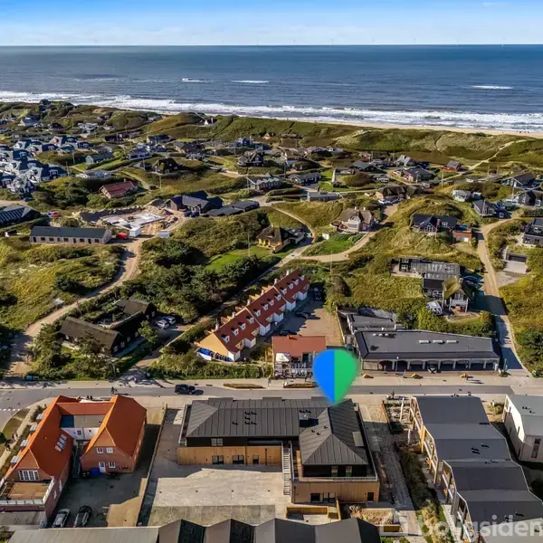 Bygninger i en kystby ligger tæt ved stranden. En farverig markør indikerer en specifik bygning. Omgivelserne har grønne områder og andre huse spredt over kuperet terræn langs kysten.