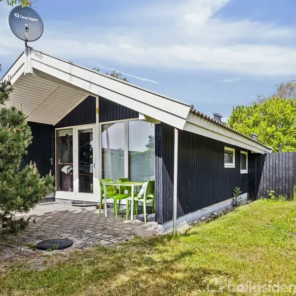 Sommerhus med sort træfacade har udestue og parabol på taget. Et sæt grønne havemøbler står på en flisebelagt terrasse. Omgivet af græsplæne, træer og blå himmel.