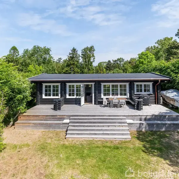 Et sort træhus står stille på en stor træterrasse omgivet af grønne buske og træer. Sollys falder på haven, der leder op til huset.