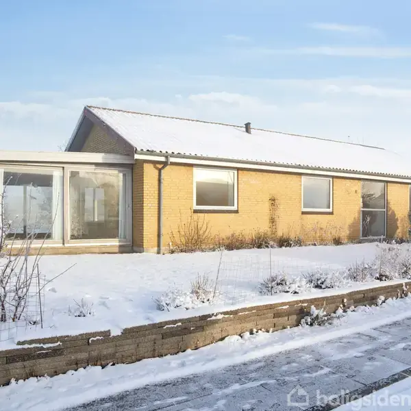 Et étplanshus står stille omgivet af en snedækket have på en klar vinterdag. Huset har gule mursten og store vinduer.