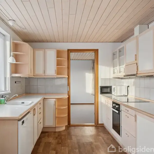 Køkken med hvide skabe og træbordplader, indeholder opvaskemaskine, vask og komfur; rummet har trægulv og træloft. Gennemsigtig vinduesgardin ved siden af vasken, døren fører til tilstødende rum.