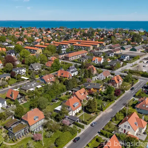 Et beboelsesområde med rødt tegltag. Husene ligger langs træfyldte gader. I baggrunden ses flere bygninger og en kystlinje med blåt hav under en klar himmel. "Boligsiden" vandmærke ses nederst.