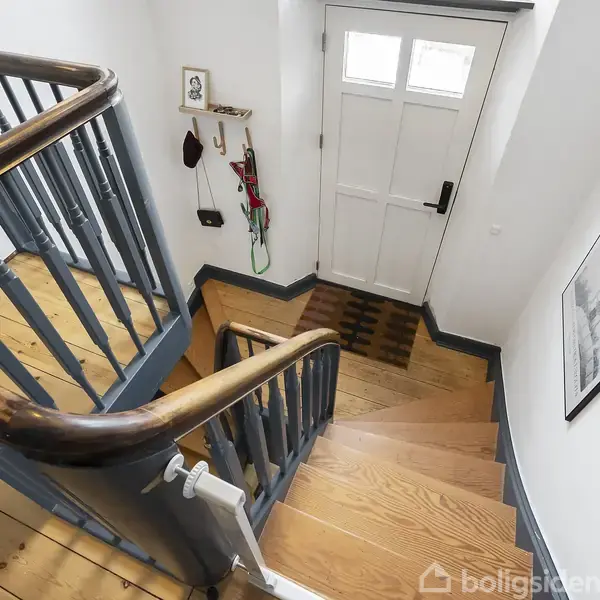 En trætrappe fører ned til en hoveddør med vinduer. På væggen hænger kroge med nøgler og en hylle med småting. Billeder pryder den hvide væg.