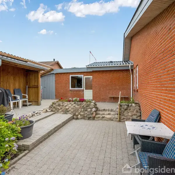 Terrasse med borde og stole ligger i et gårdhavemiljø med røde murstensbygninger. En overdækket garage er til venstre, omgivet af blomster og en lille trappe.
