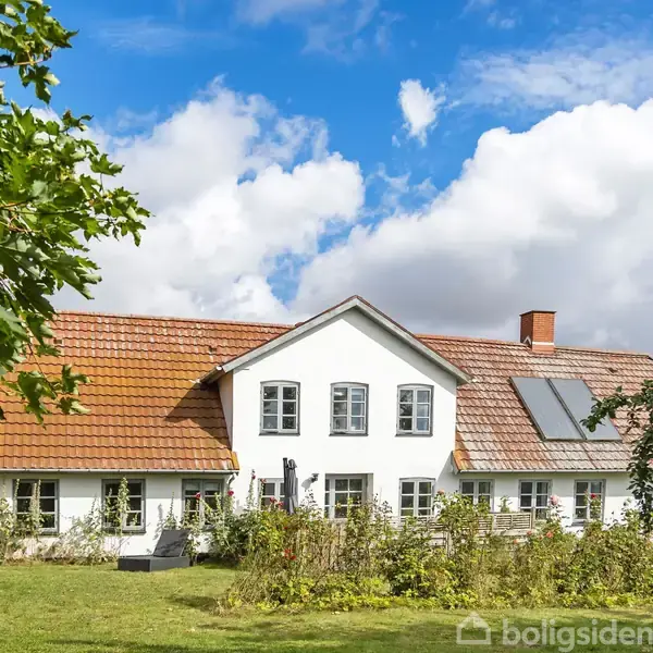 En hvid villa med rødt tegltag og solpaneler står i en frodig have. Blå himmel og skyer er i baggrunden, mens grønne træer omgiver huset.