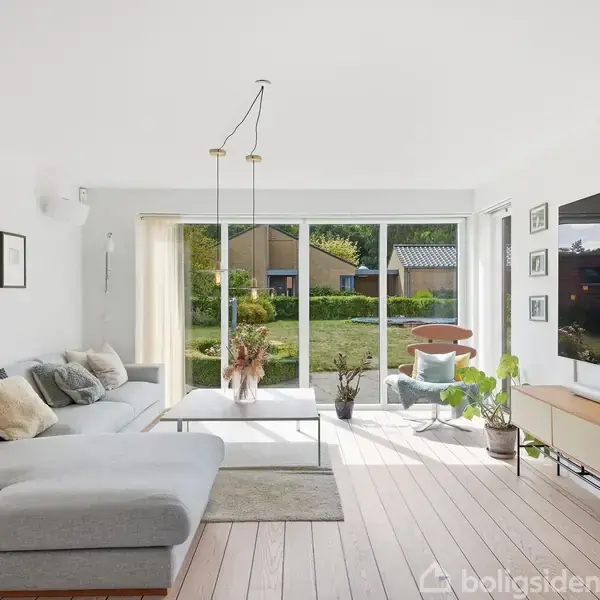 En lys stue med en stor grå sofa står foran glasdøre, der fører ud til en have. Et sofabord og flere planter skaber en hyggelig atmosfære.