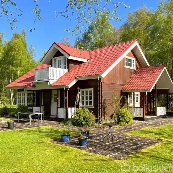 Et træhus med rødt tag står i en frodig have, omgivet af grønne træer og en blå himmel. Fliser pryder området foran huset, og der er krukker med planter.