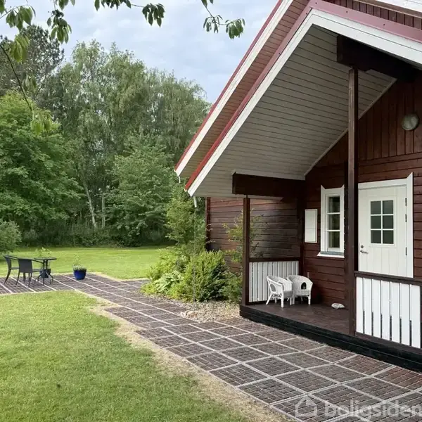 En træhytte med hvidt tag fungerer som hus, der står i en have. Ved siden af hytten er en terrasse med bord og stole, omringet af grønne træer og græsplæne.