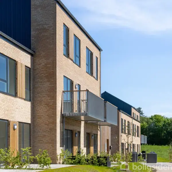 Moderne bygninger med murstensfacade står stille i et grønt område. Der er altaner og store vinduer, mens træer og græs omkranser ejendommen.