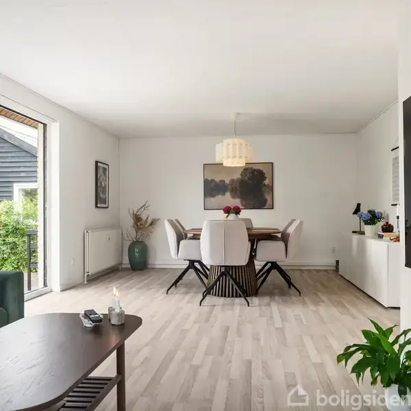En stue med et spisebord og seks stole, placeret under en hængende lampe. En grøn sofa og et sofabord står nær store vinduer med udsigt til haven. En fladskærm er monteret på væggen.