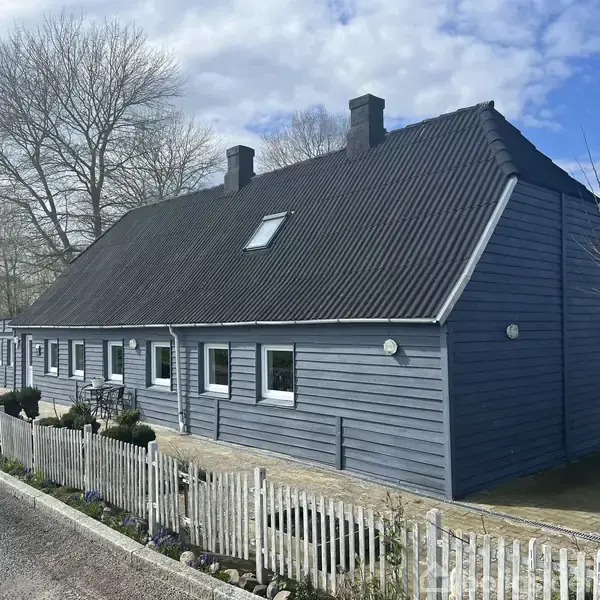 Blåt træhus med sort skråtag står i en have med hvide stakithegn. Store træer og buske omgiver huset under en blå himmel med skyer.