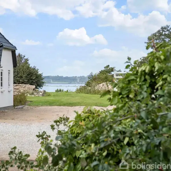 Hvidt hus står stille på grusvej, omkranset af grønne buske med udsigt til en sø i horisonten under en let skyet himmel.