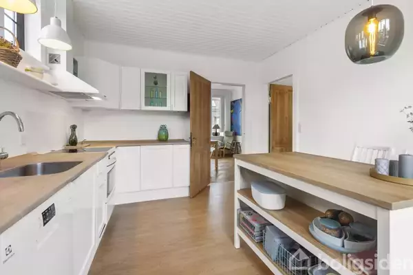 Et moderne køkken med hvide skabe og træbordplade. I midten står et åbent træbord med hylder. I baggrunden ses en åben dør til en spisestue.