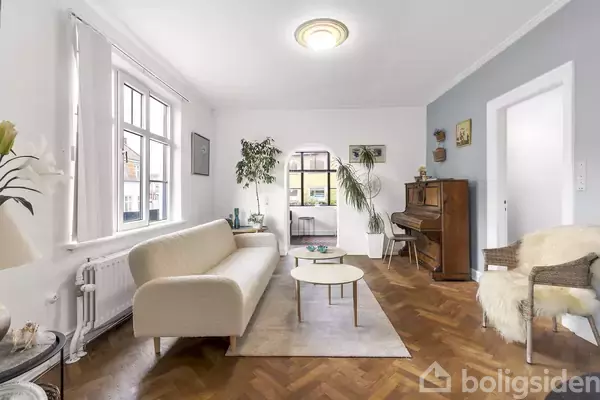 Stue med hvid sofa og rundt sofabord på et tæppe, omgivet af planter og et klaver mod en lyseblå væg. Vinduerne lader naturligt lys strømme ind.