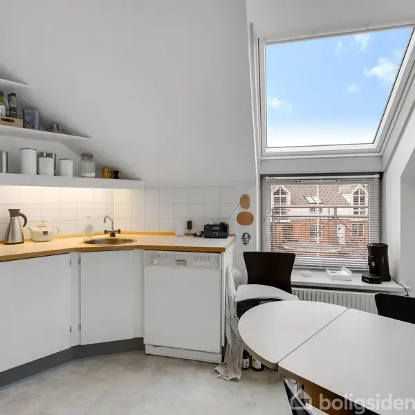 En stue med en sort lænestol, et sofabord og en brun lædersofa. Spisestue med træstole står i baggrunden. Bøger fylder hylderne, og flere lamper belyser rummet.