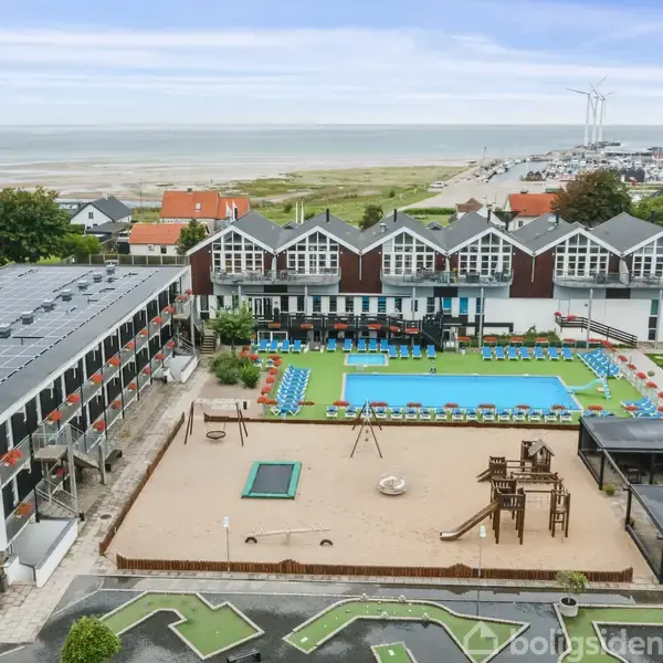 Et hotelkompleks med en centralt placeret swimmingpool, omkranset af liggestole. Der er en legeplads med sand og klatrestativer. I baggrunden findes kystlinje og vindmøller ved havet.
