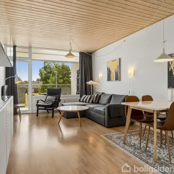 Stue indeholdende en grå sofa, et sofabord og fire spisebordsstole omkring et hvidt bord. Store vinduer åbner op mod en balkon med udsigt til grønne træer.