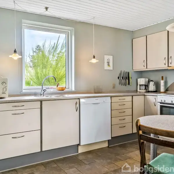 Køkken med hvide skabe, hvor en mikrobølgeovn og kaffeapparat er placeret. En opvaskemaskine står under et vindue med udsigt til grønt. Rundt spisebord kompletterer rummet.