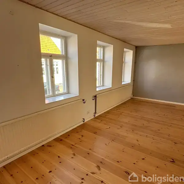 Et lyst rum med trægulv, fem vinduer langs væggen og en neutral farvetone. Rummet er tomt og godt oplyst af naturligt lys, uden møbler eller dekorationer.