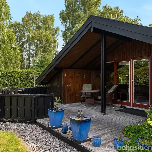 Sommerhus i træ med overdækket terrasse, placeret i en grøn have med græsplæne. Blå potteplanter står på terrassen, omgivet af buske og træer i baggrunden.