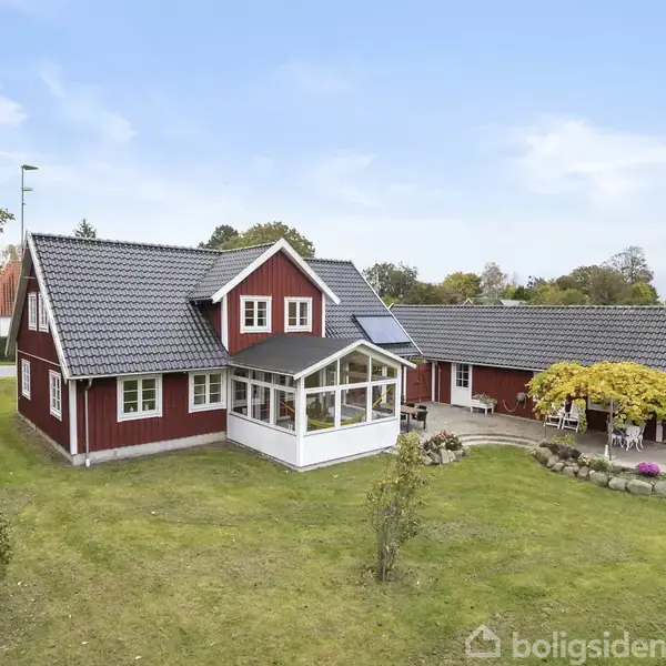 En rød villa med sort tag står i en grøn have. En terrasse med havemøbler ses ved siden af huset. Omgivet af træer og en blå himmel.