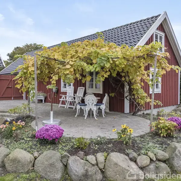 Rødt hus med hvide vinduer, hvor en terrasse overdækket af vinranker har hvide havemøbler. Omkring terrassen er der en stenbed med blomster. Græsplæne og hække i baggrunden.