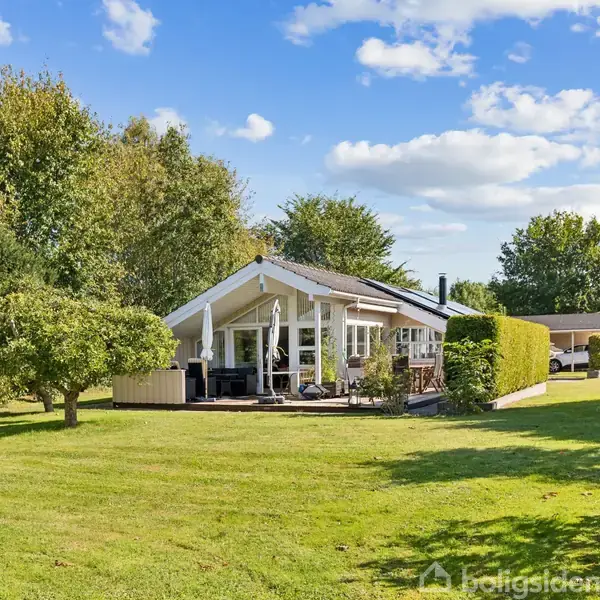 Et sommerhus står omgivet af en velplejet have med grønne buske og træer under en blå himmel med skyer. Huset har en terrasse med havemøbler.