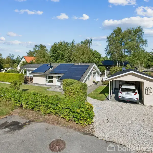 Et parcelhus med solpaneler på taget står omgivet af en grøn have med buske. Til højre er en carport med en parkeret bil. Det er solrigt og klart vejr.