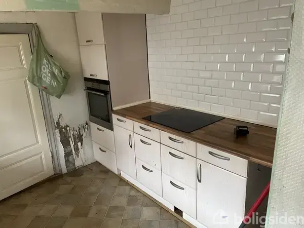 Bindingsværkshus med stråtag står i en have med en blomstrende busk til venstre. Huset har hvid facade og sorte dør- og vinduesrammer. Grå himmel i baggrunden.