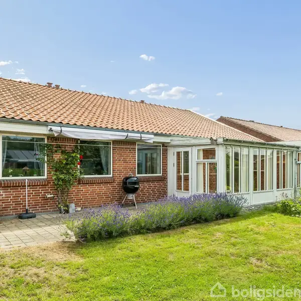 Rød murstensvilla med solarium, omkranset af grøn have. Lavendler vokser tæt på terrassen. Grill står parat ved huset. Klart, solrigt vejr med få skyer.
